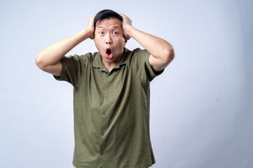 An Asian man in a casual green shirt holds his head with both hands and opens his mouth wide in...