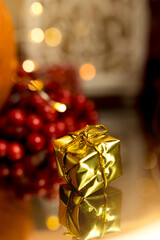 Christmas gift - Small golden parcel with golden string, among Christmas ornaments and lights. In the background a red berry. Christmas decorations and gestures of mutual memory and kindness. Vertical