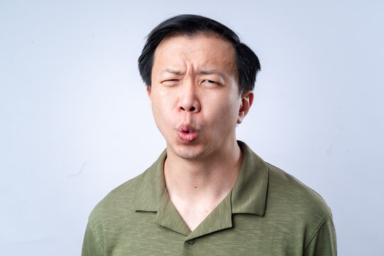 A close-up of an Asian man in a green shirt making a quirky, puckered expression with raised eyebrows and a tilted head, against a plain white background, showcasing a humorous