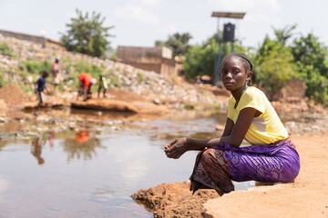 Against the backdrop of a garbage dump and dilapidated buildings, a young African woman sits on the...