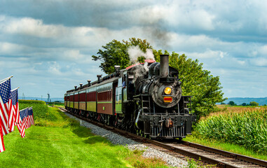 Naklejka premium A vintage steam locomotive chugs along railway tracks surrounded by lush green fields and American flags. The journey captures the charm of rural America, showcasing a beautiful day.