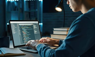 Asian male coding on laptop at desk with notebooks and coffee mug