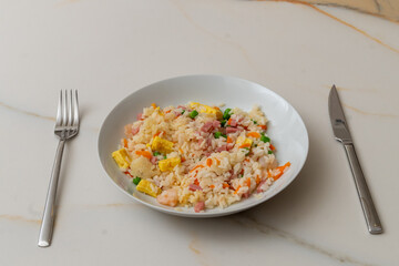 a plate of fried rice on a white marble table, with omelette, peas, ham, carrots and shrimp next to a stainless steel fork and knife