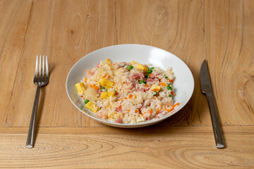 a plate of fried rice on a wood table, with omelette, peas, ham, carrots and shrimp next to a stainless steel fork and knife