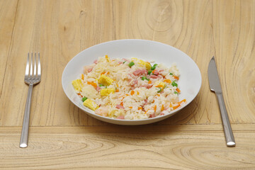 a plate of fried rice on a wood table, with omelette, peas, ham, carrots and shrimp next to a stainless steel fork and knife