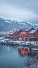 Fototapeta premium Scenic waterfront lodge with snowy mountains and warm lights reflecting in water.