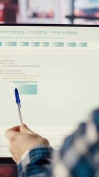 Programmer at desk typing code, developing software programs, entering commands. Man in home office fixing errors in application, implementing new features pointing to code with pen, camera B
