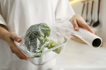 Woman putting plastic food wrap over glass container with broccoli in kitchen, closeup