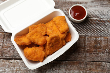 Delicious chicken nuggets in takeaway container and ketchup on wooden table, above view