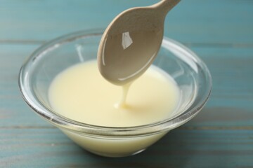 Condensed milk flowing down from spoon into bowl on light blue wooden table, closeup
