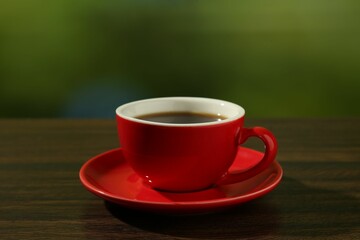 Red cup of aromatic coffee and saucer on wooden table against blurred green background, closeup