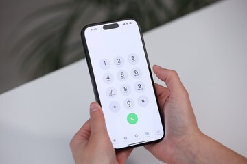 Woman holding smartphone with dialer application at white table against blurred background, closeup