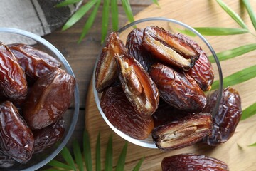 Many tasty dried dates in bowls and leaves on wooden table, flat lay
