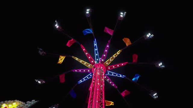 Illuminated flying chairs attraction, reaching its maximum height under a night sky, with bright lights creating a visual spectacle at the fair.