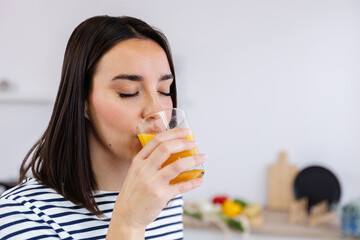 Vertical shot of young beautiful woman drinking natural orange juice in the morning in the kitchen at home. Healthy lifestyle concept