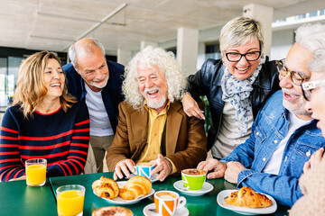 Happy group of senior people laughing and enjoying coffee at cafeteria bar. Retired generation community having fun gathered on terrace toasting hot drink. Elderly friendship lifestyle concept.