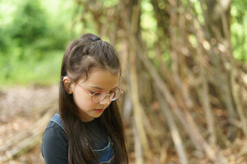 Little cute girl in denim overalls and glasses builds a house of sticks in the forest in summer. Concept of children's fun, outdoor games