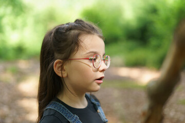 Little cute girl in denim overalls and glasses builds a house of sticks in the forest in summer. Concept of children's fun, outdoor games