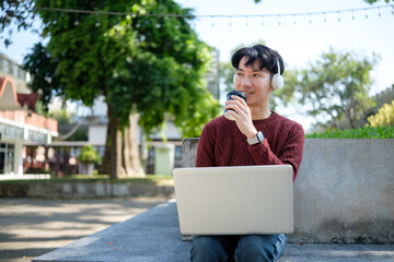 Carefree asian male student sipping coffee while working on his laptop in a campus park.