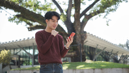 Young man wearing a maroon sweater and jeans is standing outdoors in a modern park environment.