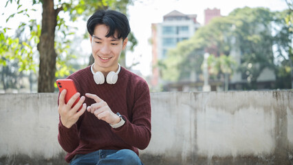 Obraz premium Happy young asian man sitting on an outdoor bench, holding smartphone and smiling.