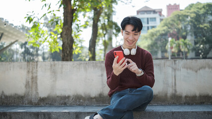 Obraz premium Happy young asian man sitting on park bench, holding smartphone and smiling.