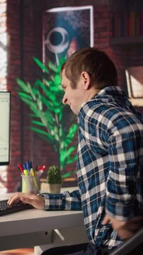 Man in home office stretches back while looking at UI on PC screen, feeling fatigued while executing programming code. IT developer feeling backpain, relieving pain, writing and testing code, camera A