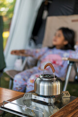 camping tent with kettle and gas stove to boil hot water with blur asian child or camper kid girl sitting on chair and outdoor camp equipment and accessory for nature holiday relax and vacation travel
