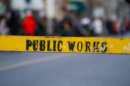 Worn, wooden ‘Public Works’ sign in front of a distant blurred crowd. Yellow, weathered sign.