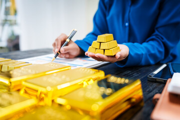 An Asian businesswoman sits at her desk analyzing gold bar prices, buying and selling gold, trading...