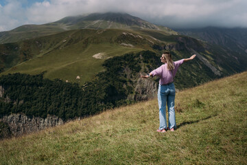 High mountains, a girl in a lilac blouse and jeans, a feeling of spaciousness and freedom. Landscapes of the Georgian military road. The girl spread her arms out to the sides