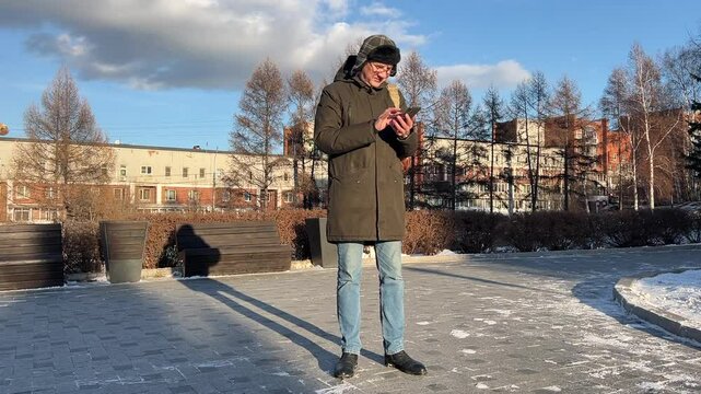 Man dressed warmly stands outside, engrossed in his smartphone in a sunny winter setting.