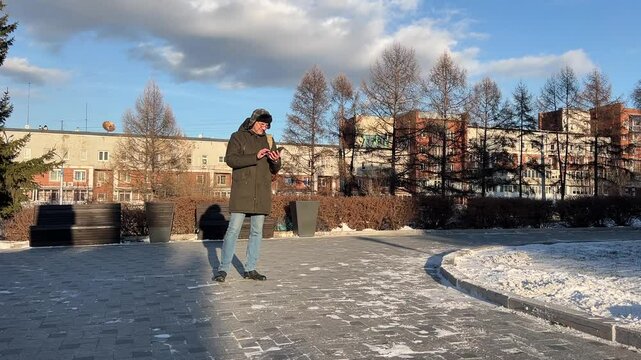 Man dressed warmly stands outside, engrossed in his smartphone in a sunny winter setting.