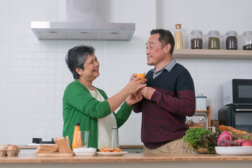 Happy Asian senior couple, wife pouring orange juice and serve her husband during cooking meal at home