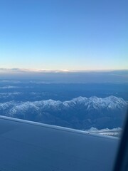 Looking out of the plane window, New Zealand's snowy mountains come into view, their jagged peaks covered in fresh snow. The pristine white slopes contrast with the deep blue sky.