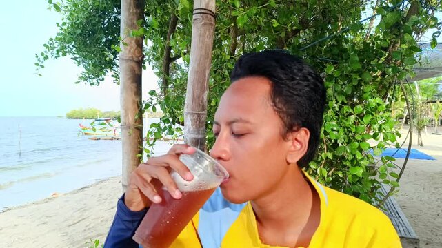 A asian guy is enjoying drinking iced tea with a beach background when summer vacation. Slow motion bit . Focus on man, 