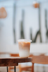 A refreshing iced coffee in a clear plastic cup, set on a wooden table. The image has a blurred background with hints of a cafe setting. Perfect for food and beverage, cafe, or lifestyle content.