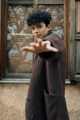 Fashionable young man in brown coat with curly hair standing by window with hand outstretched