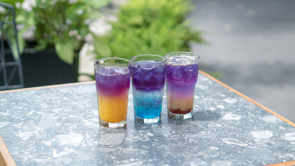 A glass of colorful two-tone drink, possibly a cocktail or refreshing beverage, sits on a textured table, outdoors. The drink has two distinct layers of color, a purple top and an orange bottom