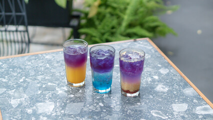 A glass of colorful two-tone drink, possibly a cocktail or refreshing beverage, sits on a textured table, outdoors. The drink has two distinct layers of color, a purple top and an orange bottom