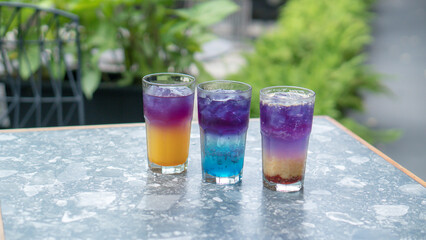 A glass of colorful two-tone drink, possibly a cocktail or refreshing beverage, sits on a textured table, outdoors. The drink has two distinct layers of color, a purple top and an orange bottom