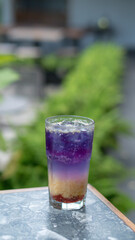 A glass of colorful two-tone drink, possibly a cocktail or refreshing beverage, sits on a textured table, outdoors. The drink has two distinct layers of color, a purple top and an orange bottom
