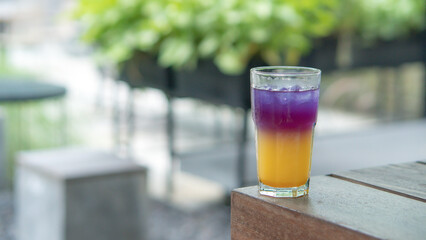A glass of colorful two-tone drink, possibly a cocktail or refreshing beverage, sits on a textured table, outdoors. The drink has two distinct layers of color, a purple top and an orange bottom