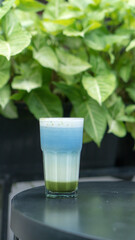 A glass of colorful two-tone drink, possibly a cocktail or refreshing beverage, sits on a textured table, outdoors. The drink has two distinct layers of color, a purple top and an orange bottom