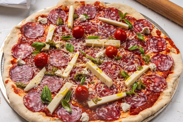 Pizza is prepared for baking. Raw pepperoni pizza with sausage, mozzarella cheese, cherry tomatoes and basil on a baking wheel