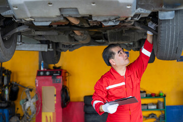 asian senior experienced mechanic business owner in uniform checking lifted vehicle in garage...