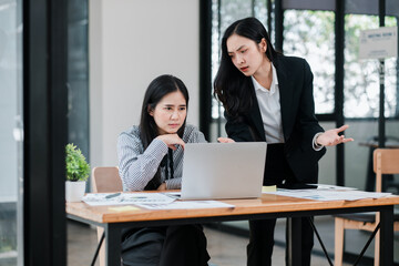 Two professional women discussing work at a desk with a laptop, showcasing teamwork and collaboration in a modern office setting.