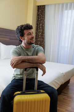 Man in his hotel room sitting on the bed with his yellow suitcase, waiting for his companion to fly to another destination or return home after the vacation.