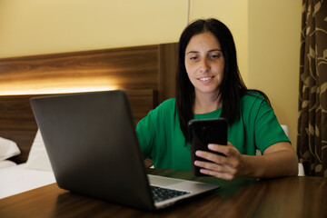 Young adult businesswoman using phone and laptop in the hotel room, she checks messages and emails to keep track of the meeting she has the next day with partners.