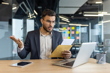 Mature businessman sits at a desk in office, reading a letter that appears to contain bad news. His...